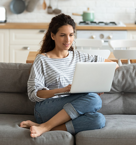 A woman sits on her couch and uses her laptop. Using a computer for long periods of time may cause digital eye strain.