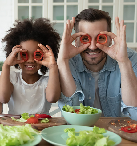A father and his daughter preparing a salad and using the pepper rings around their eyes to mimic glasses. Foods like peppers can help dry eye health!