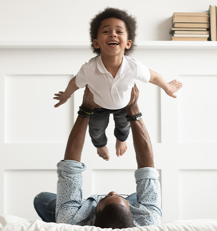 A father lifts his young son up above him while lying on the bed.
