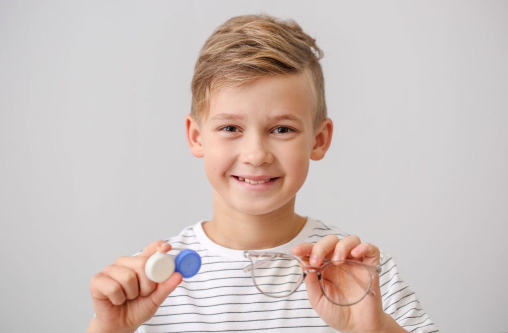 A young boy holds a pair of glasses in one hand and contact lenses in the other hand