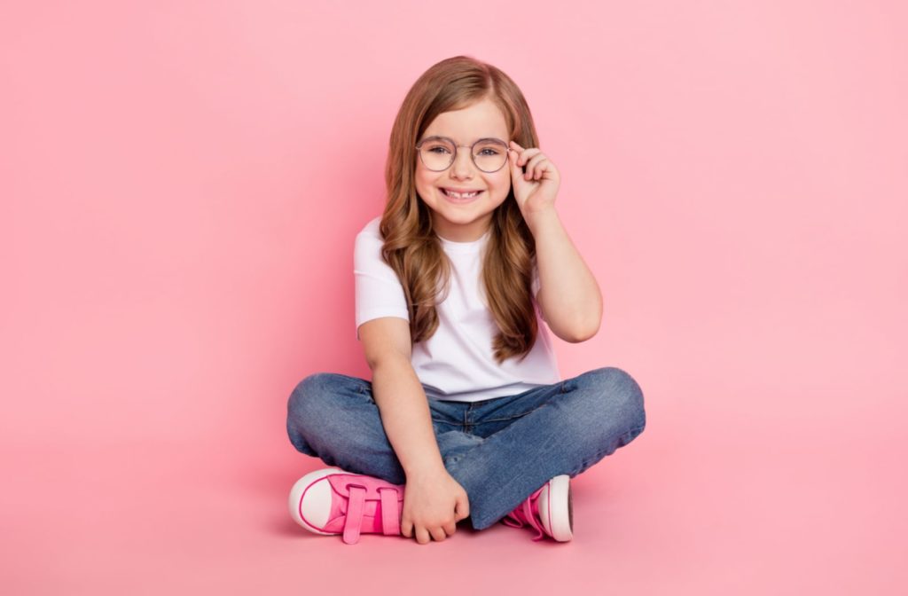 A young girl wearing glasses and sitting against a pink backdrop