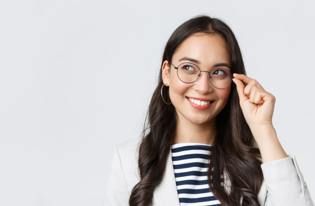 A young woman smiling and holding onto the edge of her glasses with two fingers.